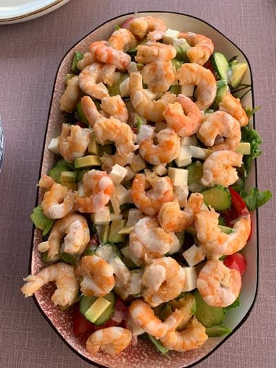 An overhead shot shows a large, oval plate filled with a shrimp salad. The salad is comprised of several ingredients, including cooked shrimp, diced avocado, cubes of white cheese, sliced cucumber, and red cherry tomatoes. The shrimp are plump and pink with a creamy dressing drizzled over them. The plate itself has a pink and white patterned rim. The salad rests on a textured, pinkish-grey tablecloth. The background shows a sliver of a white plate with a gold rim. The image is well-lit, focusing on the food. There is no indication of the city of Суроватиха, Russia, or any people present in the image.