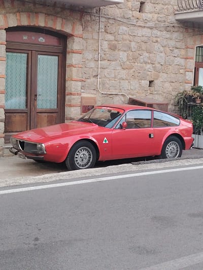 A red Alfa Romeo GT 1300 Junior coupe is parked on the side of a street in Vallo della Lucania, Italy. The car is facing left, with its front end towards the viewer. It is positioned next to a stone building with a wooden door and two windows with lace curtains. The car has a triangular emblem with a green snake on the driver's side door. The street in the foreground is asphalt with a white line marking the edge. The background shows the textured stone wall of the building and some greenery.