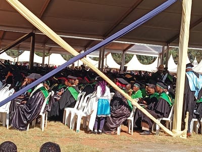 A crowd of people, mostly in graduation gowns, sit in white plastic chairs under a large tent decorated with beige and blue fabric. Many are wearing black gowns with green accents and mortarboard hats. A child with purple hair and a blue skirt stands among the seated graduates. The background features tents and trees. The event appears to be an outdoor graduation ceremony.