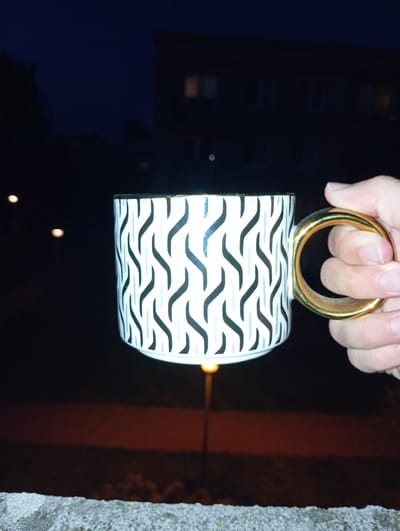 A person's hand is holding a white mug with a black wavy pattern and a gold ring handle. The mug is oriented upside down, and the hand is gripping the gold handle. The background is dark, suggesting it is nighttime. There are some blurry lights in the background, which could be from buildings or streetlights. To the right of the mug, there is a textured, gray surface, possibly a wall or a rough concrete surface. The overall impression is of a casual moment, with the mug being held up against a dark, potentially outdoor setting in Radzymin, Poland. There is no visible text in the image.