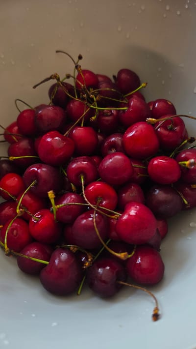 A close-up, overhead shot features a white bowl filled with fresh, dark red cherries. The cherries are glossy and appear wet, with some water droplets visible on their surfaces and in the bowl. Stems of varying lengths, some thin and brown, others thicker and green, are attached to many of the cherries, creating a tangled, organic pattern. The lighting highlights the smooth, round shapes of the fruit. The background is a plain, light-colored surface, possibly the rim of the bowl, with subtle water droplets visible. The image is an agricultural product still life.