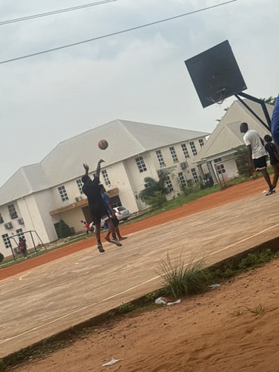 This is a media file taken in Amansee, Nigeria. The scene appears to be a basketball court with a building in the background and a dirt/grass area to the side.
The main subjects are several people, likely young men, engaged in a basketball game. One person is in mid-air, jumping to shoot or block a basketball that is visible in flight. Another person is also jumping, possibly attempting to defend the shot. Several other individuals are visible on the court, some appearing to be spectators or other players.
The building in the background is multi-story with many windows, suggesting it could be a school or dormitory. A basketball hoop with a black backboard is also visible. The court itself is marked with white lines. The ground beside the court is a mixture of dirt and grass, with some sparse vegetation and debris.
The time of day appears to be overcast, with a bright, diffused light, suggesting a cloudy day. The overall atmosphere seems to be one of casual sporting activity.
There is no visible text in the image.