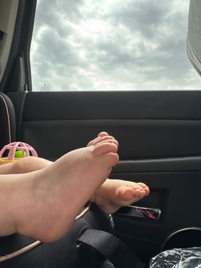 The image shows a close-up view of a baby's feet. The baby is likely in a car seat, as indicated by the presence of a car seat strap in the foreground and the car's interior door panel. A pink and green rattle is visible near the baby's leg. The background consists of a cloudy sky seen through the car window. The location is stated to be Drazhevo, Bulgaria.