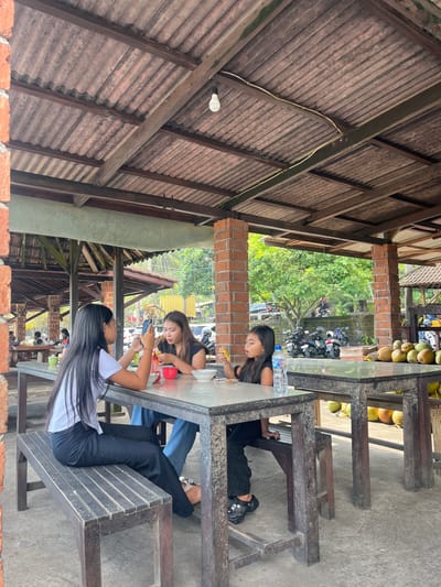 The image depicts an outdoor seating area, likely a restaurant or cafe, with a rustic, open-air design. The roof is constructed of corrugated material supported by wooden beams, and the walls appear to be made of brick. Several people are seated at a long, dark table, some of whom are engaged with their mobile phones.
In the foreground, a young woman with long, dark hair is wearing a white shirt and black pants, and is looking at her phone. To her right, another young woman in a black t-shirt is also looking at her phone. Further along the table, a third person, mostly obscured, is seated. Beyond the immediate seating area, a collection of coconuts is visible, suggesting a tropical location. In the background, several motorcycles are parked amidst lush greenery, indicating an outdoor environment. The lighting suggests it is daytime, possibly with bright sunlight. The overall atmosphere appears relaxed and casual. The location context provided is Pekutatan, Indonesia.