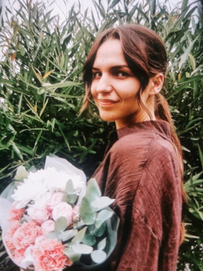 A young woman with long brown hair pulled into a ponytail stands in front of a backdrop of dense green foliage. She is wearing a brown, textured shirt and is holding a bouquet of pink and white flowers with green eucalyptus leaves. The woman is looking over her left shoulder towards the camera with a slight smile. Her facial features are visible: dark eyes, a straight nose, and full lips. The lighting suggests an outdoor setting, possibly a garden or park. There are no discernible landmarks that would indicate a specific location within the city of Serov, Russia. The image captures a portrait of the woman with a natural, slightly overcast lighting.