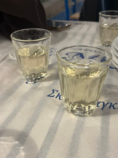 The image displays a table setting within a dining establishment in Megalochori, Greece. Three clear, vertically fluted glass tumblers, each containing a pale yellow liquid, are centrally positioned on a white paper tablecloth. The tablecloth features blue Greek lettering, partially visible as "ΣΚΕΠΑΣΤΡΟ". To the immediate right, a stack of small, white ceramic plates is visible. In the upper left background, a dark brown, indistinct object is present, likely a piece of fabric or personal item. A metallic, reflective object, possibly a knife or a mobile phone, is discernable further back on the table. No individuals are depicted, but the arrangement implies the presence of diners.