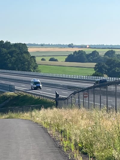 A blue and white van, possibly a police vehicle, is traveling on a highway. The van is in the middle lane of a multi-lane highway. The highway is bordered by fences and a grassy median. In the background, there are trees and fields. The sky is clear and blue. The scene appears to be in a rural or semi-rural area. The location is identified as Momberg, Germany.