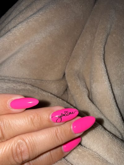 A close-up shot shows a person's hand with bright pink almond-shaped fingernails. One nail features a black cursive script that reads "you & me" with a small heart. The hand rests on a textured, tan-colored blanket. The scene is indoors, with no specific location in Fairview Township, United States of America identified.