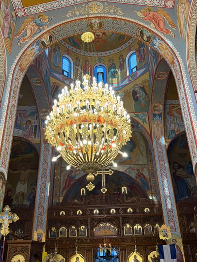 The image captures the richly decorated interior of an Eastern Orthodox church, located in Kafr Yasif, Israel. A massive, multi-tiered golden chandelier, brilliantly lit with numerous bulbs, hangs as the central focal point, illuminating the space.

The walls, arches, and vaulted ceilings of the church are entirely covered with vibrant Byzantine-style murals and icons. These elaborate paintings depict various religious figures and scenes, including angels with outstretched wings, numerous saints, the crucifixion of Jesus Christ, and Mary cradling Christ (Pietà). One prominent mural above the ornate wooden iconostasis depicts the Last Supper. The artistic style features rich colors, predominantly gold, blue, red, and earthy tones, outlined by intricate decorative borders and patterns.

Arched windows, some with blue-tinted glass, are visible higher up, allowing natural light to filter in. The heavily carved dark wooden iconostasis features multiple framed icons of saints and biblical events. Above it, a large golden cross and other golden ornaments are visible. To the left, a golden processional cross with an icon of Christ is present. A blue and white Greek flag is draped on the right side of the iconostasis. Some illegible text in Greek or Arabic script accompanies various figures in the murals. The scene conveys an atmosphere of spiritual grandeur and devotion.