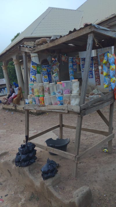 An outdoor market stall or roadside shop is depicted under a clear daytime sky. The stall is constructed from rough-hewn wooden beams and features a corrugated metal roof.
Inside the stall, a variety of products are displayed. Numerous colorful packaged snacks, such as chips and biscuits, hang from strings, forming curtains across the front and interior of the structure. Below these, several rows of clear plastic containers are stacked on wooden shelves, filled with an assortment of loose items including candies and other snacks. The branding on some packages is visible but illegible due to resolution.
In the lower-left quadrant of the image, several individuals are gathered near the stall. These include an adult woman, possibly attending the shop, and multiple children, some partially obscured. They appear engaged in daily activities, potentially shopping or socializing.
The ground surrounding the stall is an unpaved dirt or sandy surface. To the right of the wooden structure, a black plastic basin or bucket is visible, alongside several black plastic trash bags resting on the ground. A light-colored building with a patterned roof is partially visible in the upper-left background. The scene portrays a bustling, informal retail environment.