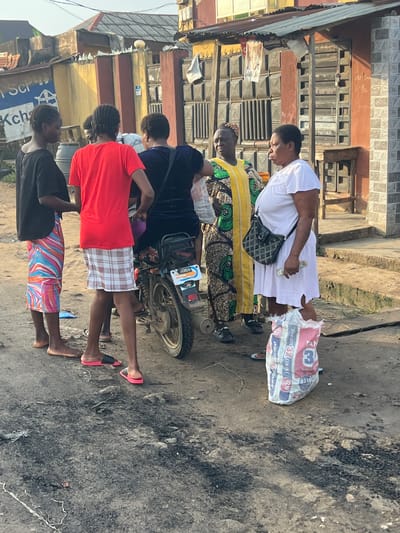 The image captures a street scene in Ijegun, Nigeria, with several people gathered near a motorcycle. A woman in a white dress stands holding cash, facing a woman in a yellow and green patterned dress. Two younger individuals, one in a black shirt and a colorful skirt, and another in a red t-shirt and plaid skirt, are also present. The ground is paved with dirt and debris, and a partially visible motorcycle with a license plate reading "BOSS" is parked nearby. In the background, buildings with corrugated metal roofs and textured walls line the street. A large bag of cement with the brand "DANGOTE 3X" is prominently placed in the foreground. The overall environment suggests a busy, everyday street scene in a Nigerian urban setting.