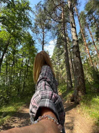 A first-person perspective shot captures a person in a forest. The subject is wearing a plaid shirt with pink and gray colors and a bracelet on their wrist. The forest setting is characterized by tall pine trees, lush green foliage, and a path winding through the trees. Sunlight filters through the canopy, creating dappled light on the forest floor. The location is specified as Portovoe, Russia.