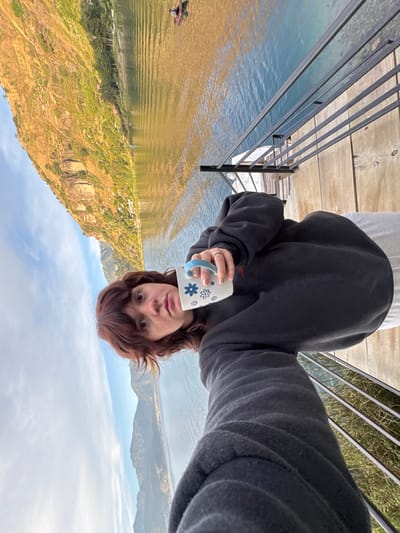 A selfie-style image captures an individual on a wooden deck in San Pablo La Laguna, Guatemala, overlooking Lake Atitlán. The person, a young adult with reddish-brown hair and bangs, is wearing a dark grey sweatshirt and light-colored lower attire. They hold a white ceramic mug with light blue handles and a blue floral design in their right hand. This hand features light pink nail polish and a silver ring on the ring finger. The individual is looking directly towards the camera.

In the foreground and mid-ground, a dark grey metal railing separates the deck from the lake. The water of Lake Atitlán displays varied tones, transitioning from yellowish-brown closer to the foreground to a deeper blue further out, with visible surface ripples. A single individual in a small boat is present on the lake in the mid-distance, positioned towards the left. The background features steep, vegetated mountains rising directly from the lake, with scattered structures visible on the slopes. The sky above is light blue with scattered white clouds.