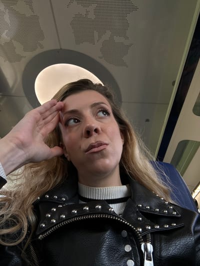 A fair-skinned female with long blonde hair is positioned in what appears to be a train carriage. She is wearing a black leather jacket with silver studs and a black and white striped sweater. Her left hand is raised to her forehead. The interior of the train is visible, including a window with a circular light fixture above it and a panel featuring a map of the world composed of small holes. The train is likely near the city of Lelystad, Netherlands.