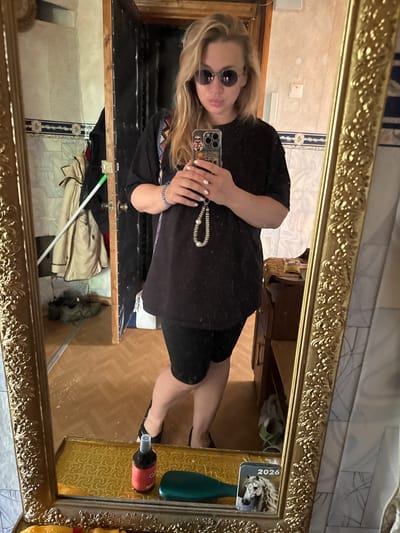 A woman with blonde hair and sunglasses takes a mirror selfie. She is wearing a black t-shirt, black shorts, and black sandals. She has a beaded necklace and a bracelet on her left wrist. She is holding a phone in her right hand. In the background, there is a door, a coat rack with jackets, and a mirror on the floor. The image appears to be taken in a residential setting, possibly an apartment in Valday, Russia.