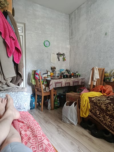 This is a candid, eye-level indoor shot of a cluttered room, likely in a living space that doubles as a kitchen or storage area, in Dovhe, Ukraine. The camera is positioned low, focusing on a person's legs and feet in the foreground, resting on a pink and white patterned blanket. The legs are bare from the knee down, with the lower legs covered by gray shorts. The person is lying on a bed or makeshift sleeping area covered in a patterned blanket.
To the left of the frame, a collection of clothing, including a bright pink towel and other garments, hangs on what appears to be a rack or a part of the doorframe. A doorway is visible behind the hanging clothes.
The midground of the image is dominated by a table covered with a patterned tablecloth, laden with various items. This includes dishes, bottles of water, cleaning supplies, and food containers, suggesting a dining or preparation area. A wooden chair is pulled up to the table, with clothing draped over its back. Behind the table, kitchen utensils are neatly organized on a wall-mounted rack. A green-faced clock hangs on the wall above the table, showing the time to be approximately 2:38.
Further into the background, to the right, is a bed or couch covered with a brown and beige patterned throw. On this surface, a bright yellow garment and several orange plastic bags are scattered. Below the bed, a cardboard box with the "GEM" logo visible on it is placed on the floor. A pair of black athletic shoes are tucked underneath the bed. The walls have a textured, grey finish, and the floor is made of light-colored wood laminate. The overall impression is one of a lived-in, somewhat disorganized but functional space. The lighting suggests it is daytime.