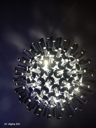 The image shows a close-up, low-angle shot of a unique, spherical light fixture. The fixture is composed of numerous metallic, cylindrical elements that radiate outwards from a central point. These elements appear to be hollow and have a reflective surface. Light emanates from within the fixture, casting a warm glow and creating a starburst effect against the dark background. The background itself is a deep, textured dark color, possibly a wall or ceiling, with subtle light patterns. The text "A1 Alpha 20+" is visible in the bottom left corner, indicating the device used for the photograph. There are no people or animals visible in the image. The scene appears to be indoors, within a residential or commercial space.