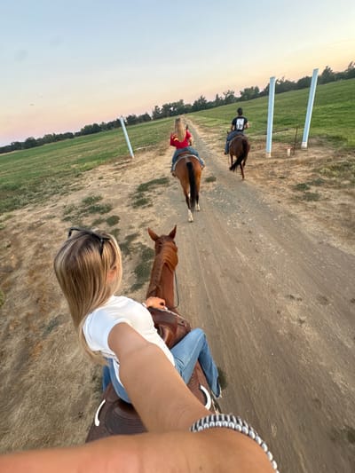 Three individuals are observed riding horses single-file down a dirt path in a rural area identified as Sloughhouse, United States, during either sunset or sunrise.
In the immediate foreground, a blonde individual, visible from the upper back, wears a white t-shirt and blue jeans, with sunglasses positioned on top of their head. This individual is seated on a brown horse equipped with a brown western saddle. Their left arm, adorned with a silver-toned bracelet featuring black segments, is extended forward, indicating a selfie-style photograph from the rider's perspective.
Directly ahead, a second individual, a blonde female, is seated on a light brown horse with a dark brown tail. She wears a red short-sleeved shirt, possibly with a yellow emblem on the back, and blue jeans, using a western saddle with decorative elements.
Further down the path, a third individual, a male with dark hair, rides a dark brown horse. He wears a black short-sleeved shirt, potentially with a white graphic on the back, and dark trousers.
The path itself is unpaved, light brown, approximately 2-3 meters wide, and appears dry with some sparse, dry grass patches. On both sides of the path, flat, green grassy fields extend outwards. Several tall, light-colored, cylindrical posts (possibly white or light blue PVC) are vertically inserted into the ground within these fields, flanking the path at various points in the mid-ground. A line of green trees forms the distant horizon. The sky exhibits a clear gradient from light blue overhead to faint orange-pink near the horizon. All three horses are moving forward in the same direction along the path.