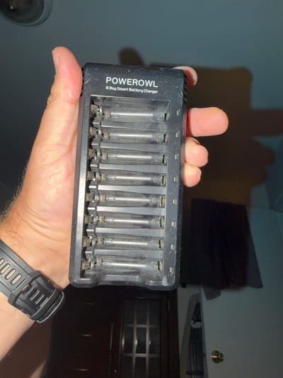 A hand in Pimmit Hills, United States of America, holds a black Powerowl 8-Bay Smart Battery Charger with multiple AA or AAA batteries inserted. The charger is mostly full, with batteries occupying most of the slots. The hand is positioned on the left side of the image, supporting the charger. The background is dark and out of focus, suggesting an indoor, dimly lit environment. The charger appears to be made of plastic and shows some signs of dust or wear in the battery slots. A watch is visible on the wrist of the hand holding the charger.