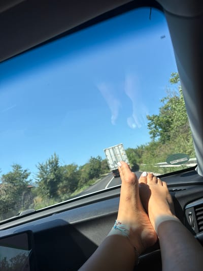 This image depicts a pair of bare feet, painted with white nail polish, resting on the dashboard of a car. The feet are positioned in the foreground and occupy the lower right portion of the frame. Beyond the dashboard, a clear blue sky is visible through the car's windshield, with some trees and a road in the middle distance. A white truck trailer is also visible on the road. The overall impression is that of a person traveling by car on a sunny day. The image does not contain enough information to determine the specific location within Medovnitsa, Bulgaria.