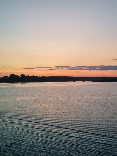 This image depicts a serene waterscape at dusk, likely in the vicinity of Nikol'skaya Gora, Russia. The sky transitions from a pale blue at the top to a warm orange and pink hue near the horizon, indicating the setting sun. A dark, silhouetted treeline forms a natural border between the sky and the water, suggesting a wooded coastline. The water itself is calm, with subtle ripples creating a textured surface. In the foreground, distinct wave patterns are visible, suggesting movement, possibly from a boat or the natural flow of the water. There are no visible people or buildings in the frame. The overall atmosphere is peaceful and natural.