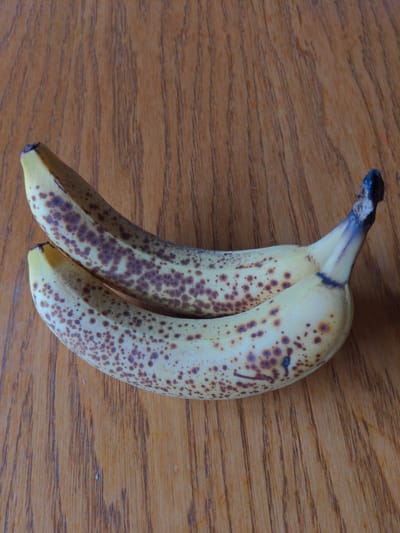 The image shows two ripe bananas with brown spots lying on a wooden surface. The wooden surface has a prominent grain pattern. The bananas are positioned diagonally across the frame, with their stems pointing towards the top right. One banana is slightly above and behind the other. The lighting suggests a natural light source. The scene is devoid of any urban elements and therefore cannot be located in Magherafelt, United Kingdom.