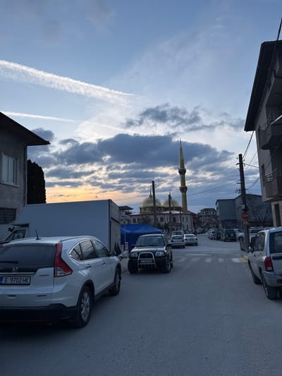 The image displays an urban street scene in Breznitsa, Bulgaria, captured during twilight under a sky with scattered grey clouds, prominent contrails, and a low sun casting an orange glow near the horizon.
In the middle ground, the central feature is a mosque characterized by two prominent golden domes and a tall, slender minaret, which has a green band and culminates in a golden, pointed finial.
The asphalt street extends into the distance, flanked by buildings and parked vehicles. In the foreground on the left, a white Honda CR-V SUV, bearing Bulgarian license plate E 9702 HK, is parked. Behind it, a large, rectangular white utility structure or vehicle body and a blue tent structure are visible. Further down the left side, a dark-colored SUV, equipped with a front bull bar and displaying Bulgarian license plate E 3477 HB, is parked.
On the right side of the street, a silver SUV is parked in the midground. A black utility pole with multiple overhead wires is present on the right, positioned near a red octagonal 'STOP' sign. Buildings with various facades and heights, including a two-story structure on the immediate left and a multi-story building with balconies on the immediate right, frame the street. A pedestrian zebra crossing is painted on the asphalt surface in the mid-foreground. No individuals are visible in the scene.