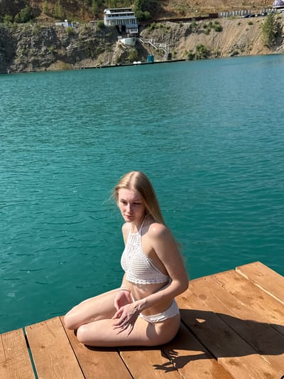 A young woman with blonde hair is sitting on a wooden pier next to a body of turquoise water. She is wearing a white crocheted bikini top and white bikini bottoms. The water is calm, and the pier is made of weathered wooden planks. In the background, there is a steep, rocky slope with some vegetation and a building on top. The sky is clear and blue. The location is identified as Karyer Izvestnyak, Russia.