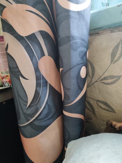 The image shows a close-up view of a rolled-up textile, likely a curtain or a piece of fabric, with a floral or abstract pattern. The pattern consists of large, stylized leaf-like shapes in shades of peach and dark teal, with lighter grey and white accents. The fabric is creased and folded, creating shadows and highlighting the texture. To the right of the fabric, a section of a wall or another textile with a faint, grey leaf pattern is visible. The background is out of focus, but hints of a shelf with some items, possibly a bottle with a label showing the number "90," can be seen on the left. The overall impression is that of an indoor setting, possibly a room in an apartment in the city of Pervomaisky, Russia, where this textile is part of the decor.