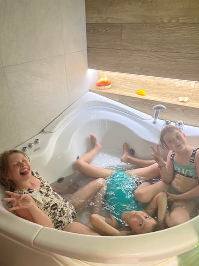 The image shows four young people inside a white bathtub. The bathtub is positioned in a room with light-colored tile flooring and wooden-style wall paneling. The people are in the water; one is lying with her head near the edge, laughing, and wearing a floral pattern top. The others are seated or positioned within the tub, wearing swimsuits. Location is Новая Чернушка, Russia.