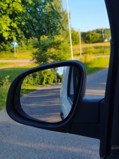 The image shows a view from inside a vehicle looking into the side mirror. The mirror reflects a road winding through a green, treed landscape under a clear blue sky. The road is paved and shows tire marks and the reflection of sunlight. In the foreground, the black frame of the side mirror and a portion of the car's door pillar are visible. To the left of the mirror, green foliage and a blue signpost are visible outside the car. The overall scene suggests a drive through a rural or suburban area, likely during daytime given the bright sunlight. Based on its characteristic landscape and vegetation, this scene is consistent with the environment found in Aizpute, Latvia, a town known for its scenic rural surroundings.