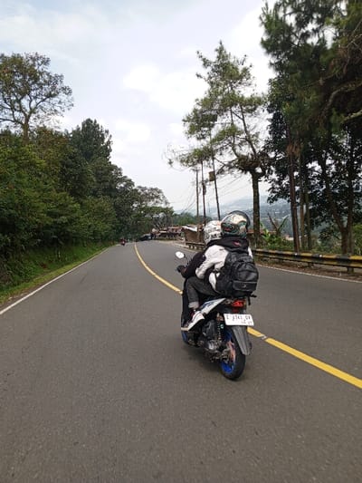Two individuals are observed riding a blue and grey scooter-type motorcycle away from the viewer on an asphalt road in Ciloto, Indonesia. The driver wears a dark jacket and a multi-colored helmet, while the passenger, seated behind, wears a white hooded jacket, a black backpack, and a white helmet with a dark visor. The motorcycle bears a license plate "Z 4741 GV 06-27" on its rear fender. The road features a double solid yellow centerline and a white solid line on its left edge, curving gently to the left. Dense green foliage and tall trees line both sides of the road. A metal guardrail is present on the right side, beyond which multiple utility poles with overhead wires and several pine-like trees are visible. Further down the road on the right, a cluster of small structures with corrugated metal roofs can be discerned. The background reveals distant hilly terrain under an overcast sky.