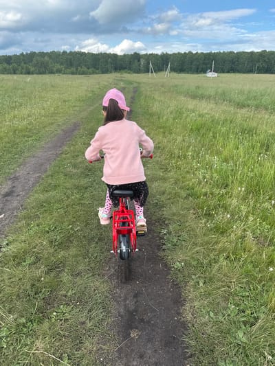 A young girl, wearing a pink sweater, a pink baseball cap, and patterned leggings, sits on a red bicycle. She is on a dirt path surrounded by tall green grass. The path appears to be in a rural area, possibly a field or meadow. In the background, there is a dense line of trees and a cloudy sky with blue patches. The overall scene suggests an outdoor activity on a pleasant day. The girl appears to be actively riding her bicycle.
