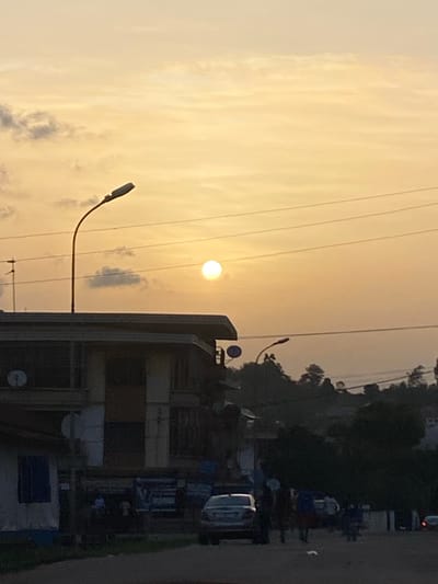 The image shows a view in Abofifia, Nigeria, likely during sunset or sunrise, given the golden hue in the sky. Overhead power lines are visible against the sky. In the middle ground, there is a building with a satellite dish mounted on it. A street lamp extends into the sky. On the right side of the frame, there are parked vehicles and indistinct figures of people, possibly a market area or a roadside gathering. Trees and vegetation are visible in the background. The overall scene appears to be a typical urban landscape in Abofifia during a time of day with low sun angle.