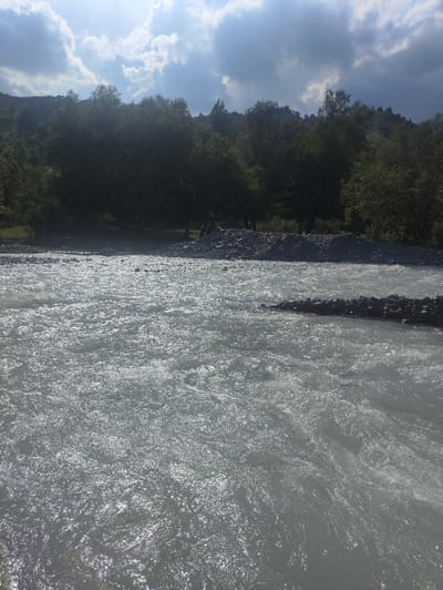 The image depicts a wide, fast-flowing river in Kashka-Suu, Kyrgyzstan, under a partly cloudy sky. The water is a milky, light gray color and appears turbulent, with numerous ripples and whitecaps reflecting sunlight. Along the near bank of the river, a barrier constructed from dark stones is visible, extending into the water. The opposite bank is lined with a dense forest of dark green trees, beyond which rises a hillside with sparse vegetation and rocky outcrops. The sky above is a mix of bright blue and heavy, dark gray clouds, with sunlight breaking through in places. The overall impression is of a natural, rugged landscape.