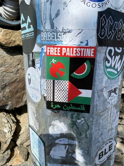 The image displays a metallic utility pole, heavily covered with numerous stickers, located in Corniglia, Italy. The pole exhibits signs of wear, including scratches, fading, and remnants of torn paper from older stickers.
Centrally affixed is a large rectangular sticker with a red band at the top displaying "FREE PALESTINE" in white capital letters. Below this, the sticker is divided into four quadrants: the top-left features a red dove with an olive branch on a green background; the top-right shows a slice of watermelon on a black background; the bottom-left depicts a black and white keffiyeh pattern; and the bottom-right presents the Palestinian flag (black, white, green horizontal stripes with a red triangle). A black strip at the bottom of this sticker contains green Arabic text, translating to "Palestine is free."
Surrounding this central sticker are various other decals, including a prominent "AC/DC" logo, an "SVW" logo, "UMO COCCO OUTDOOR EXPERIENCES," "BABELSHOP," "ITINARRANDO" with an owl graphic, "TOPLINE Feat Tiger H," and "BLE Best Life Ever Flagstaff - Arizona." A QR code sticker for "TREKKING4LIFE.PT" is visible lower on the pole.
To the left of the pole, a rough-hewn stone wall or rock formation, composed of various brown and grey stones, is partially visible. A white corrugated pipe or conduit is seen behind the upper left portion of the pole. The scene is illuminated by daylight, casting a shadow on the lower right section of the pole. No people or overt actions are depicted.