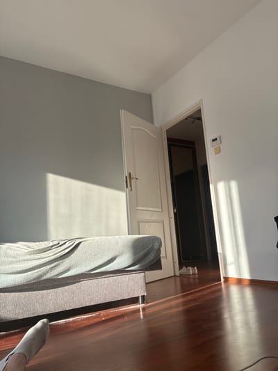 A room features white walls transitioning to gray. A light-colored door hangs open from a ceiling hatch revealing a dark-screen television. Sunlight casts long shadows. A grey sofa with a blanket sits adjacent to the wall. A person's foot is visible in the lower right corner of the image. A wooden floor is on the right, aligned with the ceiling hatch.