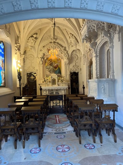 A view of a church interior showcasing intricate architectural details. Elaborate, white-colored carvings adorn the walls and ceiling. Pews made of dark wood line the patterned mosaic floor. A central painting of the Virgin Mary and baby Jesus is illuminated by a golden light. Other smaller paintings, sculptures, and decorative objects are visible along the walls. The overall scene conveys an atmosphere of solemnity.