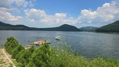 The image shows a wide, calm lake with forested hills in the background under a bright blue sky with fluffy white clouds. In the foreground, a paved path leads down to the water's edge, bordered by lush green pine trees and wild foliage. A wooden dock extends into the lake, with two small boats tied up. One boat is white with a blue trim, and a larger, white motorboat is anchored further out. The scene appears to be located at the Asparuhovo dam, near the town of Asparuhovo, Bulgaria, a popular spot for recreation and scenic views. There are no people visible in the image.
