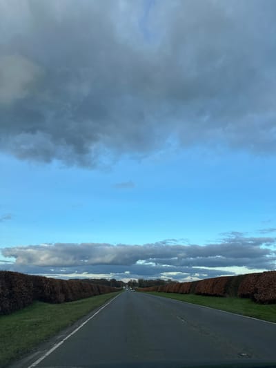 A road stretches into the distance, flanked by green grass and tall hedges on either side. The gray asphalt contrasts with the verdant surroundings. Above, the sky is split between a clear blue and a gathering storm of dark, heavy clouds. The ominous clouds dominate the left side, while the brighter blue sky fades towards the horizon, meeting the road's vanishing point. The scene suggests a blend of calm and impending weather.