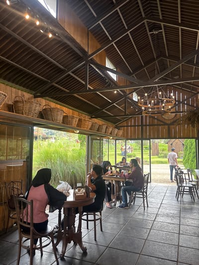 A unique perspective inside a restaurant with a high, exposed-beam ceiling featuring a chandelier and decorative baskets. People are seated at tables, some looking out through large windows onto a green, grassy area. The interior is dimly lit, contrasting with the brighter outdoor view. The arrangement of tables and chairs creates a spacious yet intimate dining atmosphere.