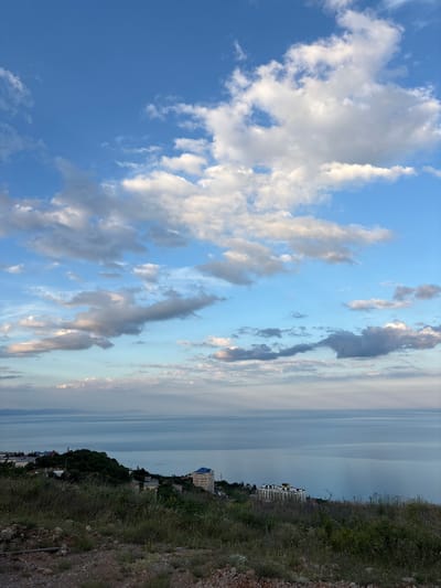 This is a landscape image of a coastal scene with a focus on the sky and the distant sea. The image is oriented vertically. The sky is a prominent feature, filled with a mix of white and gray clouds against a bright blue background, suggesting fair weather. Towards the right side of the image, the sky transitions to a hazy, lighter tone as it meets the horizon.

The sea stretches out in the background, appearing calm and reflecting the muted colors of the sky. Along the coastline, there is a green, tree-covered hillside dotted with buildings. One notable building is a multi-story structure with a light-colored facade and a dark blue roof. Further down the coast, other buildings and structures are visible, nestled amongst the trees and on the slopes. The foreground on the right side of the image is a rocky, overgrown area with sparse vegetation and dry grass, indicating a natural, untamed terrain.

The lighting suggests it is either early morning or late afternoon, given the soft quality of light and the presence of clouds. There is no discernible activity or event taking place; it appears to be a still, observational shot of the natural environment. No text is visible in the image. The scene evokes a sense of tranquility and the vastness of nature.