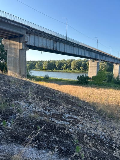 The image depicts a long, multi-span concrete and steel bridge crossing a wide, calm river. The bridge structure appears aged, with visible rust on its metal components and some wear on the concrete pillars. The riverbanks are lined with dense green trees, creating a natural backdrop to the industrial infrastructure. The foreground features a dry, grassy slope with some exposed soil and scattered debris, illuminated by the warm light of what appears to be late afternoon or early morning sun. The sky above is clear and blue, indicating fair weather. The scene is devoid of any people or vehicles, suggesting a quiet, possibly rural or undeveloped area within or near Лунненскі сельскі Савет, Belarus.