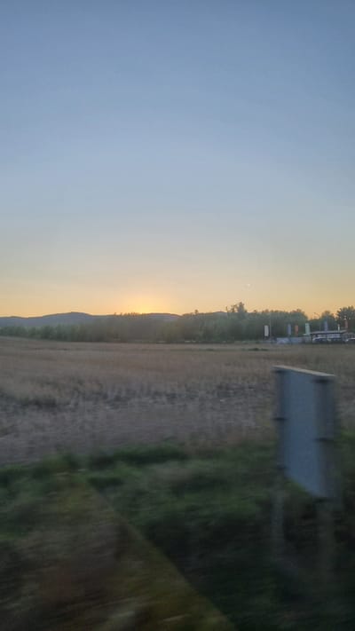 The image depicts a rural landscape at dusk, possibly near Nicolae Bălcescu, Romania. In the foreground, a blurred field of dry grass is visible, with patches of green vegetation in the lower left and right corners, also affected by motion blur. A grey, rectangular object, possibly a signpost or utility box, stands to the right of the frame, similarly blurred.

The middle ground features a harvested agricultural field with dry stubble. Beyond this, a line of trees forms a dense forest, with some lighter-colored foliage suggesting deciduous trees. In the distance, rolling hills are silhouetted against the sky.

The sky itself transitions from a pale blue at the top to a warm yellow-orange hue near the horizon, indicating the setting sun. A faint crescent moon is visible in the sky above the treeline. On the right side of the image, behind the trees, a low-lying structure with a multi-colored canopy or flags and what appear to be vehicles or sheds is discernible. The overall impression is one of a serene, rural evening.