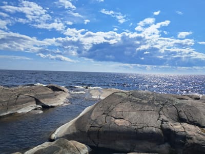 This is a landscape photograph of a rocky shoreline on a sunny day. The image features a clear blue sky dotted with fluffy white clouds. The sun reflects brightly off the surface of a dark blue body of water, likely a lake or sea, which stretches to the horizon. In the foreground, large, weathered grey rocks, some partially submerged in the water, dominate the scene. The rocks exhibit a rough texture with visible cracks and striations. The water is calm with small ripples near the shore and where it meets the rocks. The scene appears to be a natural, undeveloped area, devoid of any human presence or man-made structures. The location is Tervu, Russia.