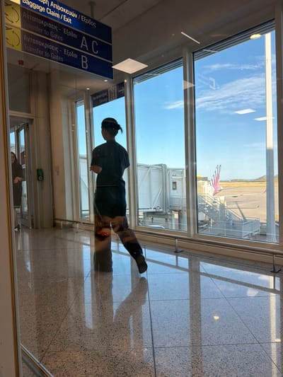 The image appears to be taken from inside an airport in Spata-Loutsa Municipal Unit, Greece. The viewpoint is through a window, which is reflecting the interior scene. Outside the window, an airplane is visible, along with an airport boarding bridge. The tail of the plane has the American flag colors. Inside the airport terminal, a person is walking by the window. The floor is tiled. A sign is visible indicating "Baggage Claim/Exit" and "Transfer to Gates" in both Greek and English, along with references to gates A, B, and C. There is also an elevator sign. The sky is clear with some clouds.
