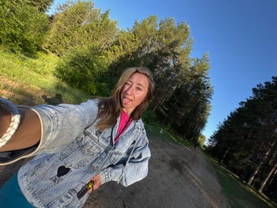 A young to middle-aged woman is depicted in a selfie, winking with her right eye and sticking out her tongue, indicating a playful expression. Her hair is long, wavy, and brown, with sunlight highlighting its texture. She is wearing a light-wash denim jacket adorned with a black heart-shaped patch on the left chest, featuring a white, stylized 'A' with a small heart. Beneath the jacket, a vibrant pink top is visible. A white, possibly woven or braided, bracelet is on her left wrist, and she wears teal-colored pants or leggings. In her right hand, she holds a yellow and black pet grooming tool, resembling a deshedding brush.

The scene is an outdoor environment, characterized by a wide dirt or gravel path that recedes diagonally into the background. Dense green foliage, primarily coniferous trees (pines) and some deciduous trees, flank both sides of the path, creating a natural forest setting. The trees on the left side appear to be on a slight incline. The sky above is clear and bright blue, suggesting fair weather. The sunlight, appearing to come from the upper right, casts long shadows and illuminates the scene, indicating either early morning or late afternoon. In the far distance along the path, a small, dark, human-like figure is faintly visible. The setting is consistent with a natural forested area in Ust'-Silayka, Russia.