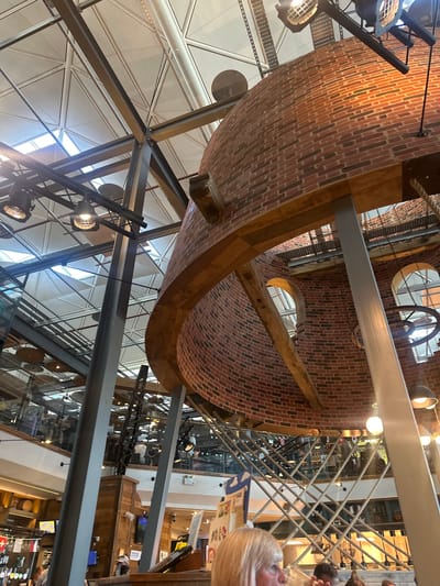 This is a high-angle, interior shot of a multi-level building with exposed brickwork and a complex metal support structure. The main subject is a large, cylindrical brick structure, possibly a silo or tower, with visible wooden beams and metal framing. Several circular windows are cut into the brickwork. The building appears to have an open-plan design with multiple floors visible, including a balcony area with rope railings.
Several people are present on the different levels, appearing to be patrons of a restaurant or bar. They are mostly out of focus and seen from behind or in profile. The atmosphere suggests a lively indoor environment, possibly a brewery or a venue with a rustic industrial aesthetic. The lighting appears to be artificial, with spotlights and overhead lamps visible.
The setting suggests a commercial establishment, likely a pub or restaurant, given the presence of what looks like a bar area with bottles on shelves in the background, and people socializing. There are some signs visible in the background, but the text is illegible. The time of day is indeterminate due to the indoor setting and artificial lighting. The overall impression is one of a well-lit, busy public space with a distinctive architectural style.