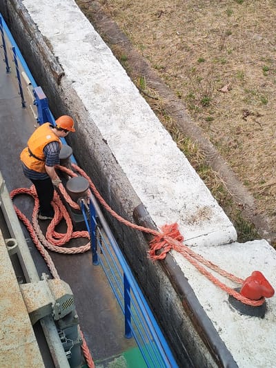 A high-angle shot shows a worker on a vessel tying it to a bollard on a quay. The worker, wearing a safety helmet and vest, is handling a thick, braided rope. The rope leads from the vessel to the red bollard, which is mounted on a concrete quay. The quay separates the vessel from a grassy area. The setting appears to be Alekseevskoe, Russia.