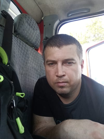 A mid-shot, selfie-style photograph shows a man inside a vehicle. The man is wearing a black t-shirt and has short, dark hair with some gray. He has a light beard and appears to be looking directly at the camera with a neutral expression. To the left of the man is a black backpack with bright green zipper pulls. Behind the man, the interior of the vehicle is visible, including a gray seat with a patterned upholstery and a headrest. The open passenger door reveals a bright, slightly blurred outdoor scene with green foliage. The lighting suggests it is daytime. The location is identified as Svémyslice, Czechia.